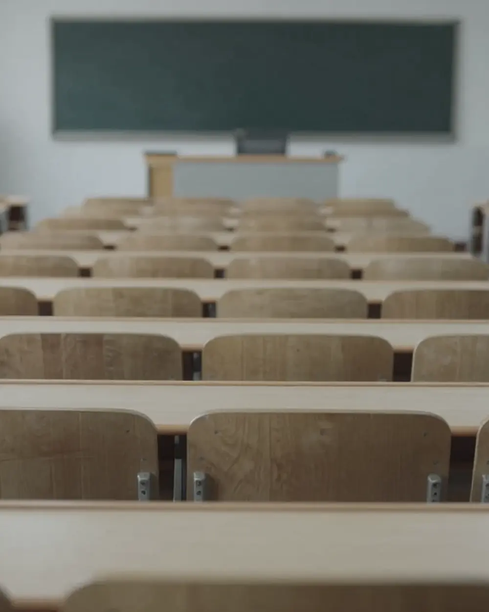 An empty tiered university lecture hall seen from the back row, rows of fold-down desks running down to a blackboard.